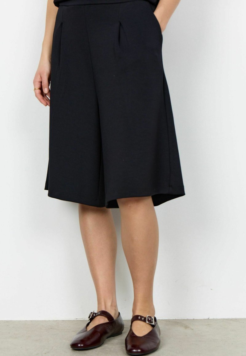 Femme portant un short large noir mi-genou avec des poches latérales et des chaussures marron foncé à boucle, debout, mains dans les poches.