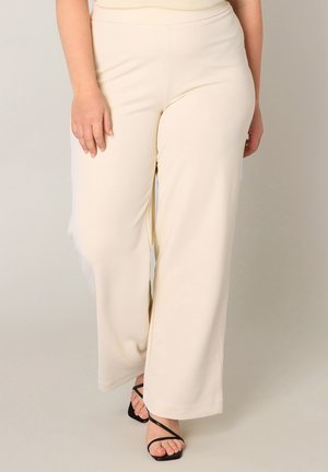 Vrouw draagt crème wijde broekspijpen en zwarte sandalen met hak en bandjes, staande tegen een effen lichte achtergrond.