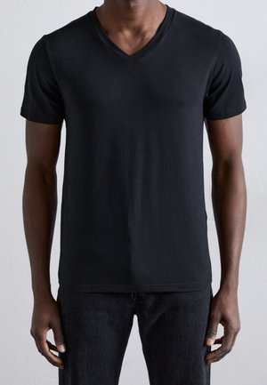 Homme portant un t-shirt noir uni à manches courtes et col en V ainsi qu'un pantalon noir, debout devant un fond clair uni.