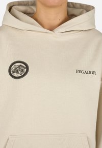 Béžová mikina s klokankou. Na prednej strane sa nachádza kruhová grafika domu a text "PEGADOR" v čiernom prešívaní.