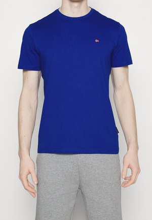 Mand iført en ensfarvet kongeblå t-shirt med korte ærmer og et lille flaglogo på brystet samt lysegrå joggingbukser mod en hvid baggrund.