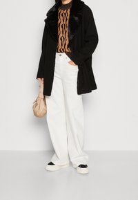 Svart kappa med fake pälskrage, brun mönstrad tröja, vita vida jeans och svartvita sneakers. Beige väska i handen.