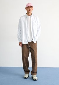 Camisa com riscas azuis claras e brancas, T-shirt branca, calças castanhas, gorro rosa e sapatilhas cinzentas chunky. Design minimalista com um corte descontraído.