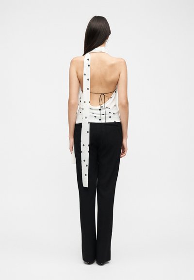 Femme aux longs cheveux foncés, de dos, portant un haut dos nu blanc à pois, associé à un pantalon noir à jambes droites.