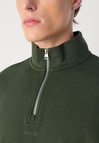 Pullover verde scuro a maniche lunghe con colletto alto, dotato di zip argentata e logo "HUGO" sul davanti. Tessuto di texture liscia.