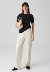 Zwart geribbelde t-shirt, crèmekleurige wijde broek, zwarte platte schoenen met strikken, stro-handtas en zwarte pet. Minimalistisch ontwerp met neutrale kleuren.