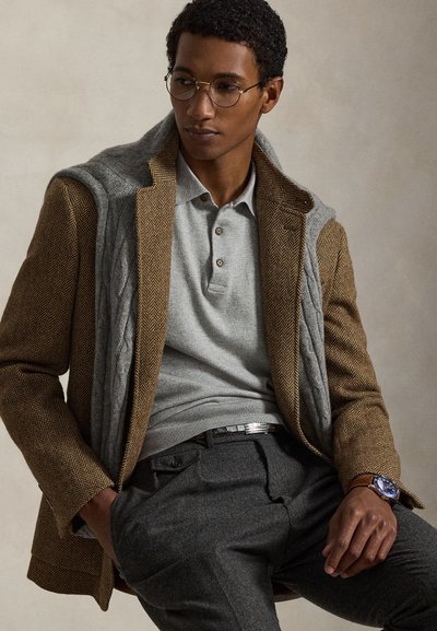 Blazer marrone a motivo pied de poule abbinato a un gilet grigio a trecce. Polo grigia, pantaloni grigio scuro e orologio con quadrante blu.