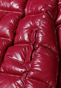 Czerwona kurtka puchowa z błyszczącą powierzchnią, z pikowanymi sekcjami i widocznym szyciem, tworzącymi teksturowany, wypchany wygląd.