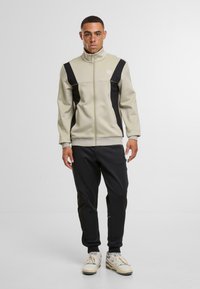 Světlý béžový zip-up svetr s černými akcenty, s vysokým límcem a logem. V kombinaci s černými tepláky a béžovými teniskami. Hladká textura.