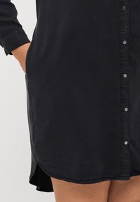 Black, button-up dress featuring a soft texture, long sleeves, side pockets, a curved hem, and silver-toned buttons along the front.