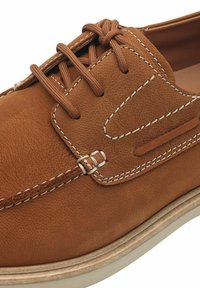 Brun lädersko med texturerad finish, försedd med bruna snören, kontrasterande vit sömnad och en krämfärgad gummisula.