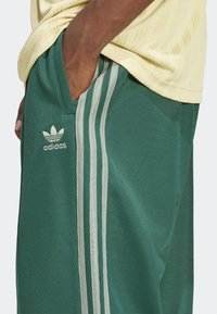 Gröna sportbyxor med strukturerat tyg, med en sidoficka och tre vertikala vita ränder. Adidaslogotyp i guld på vänster ben.