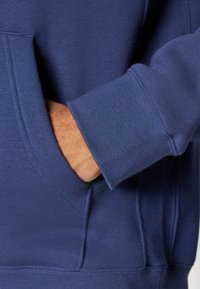 Marineblå hettegenser med frontlomme, ribbede mansjetter og myk tekstur. Synlig sømdetalj fremhever det overordnede designet.