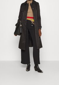 Abrigo negro de trench con un acabado suave y brillante, combinado con un sweater de cuello alto de canalé beige, pantalones anchos negros y botas de tobillo. Incluye un bolso.