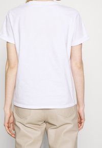 Vit t-shirt med kort ärm i bomull, med rund halsringning och avslappnad passform, visad från ryggen tillsammans med ljusbruna byxor.