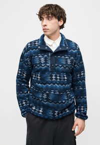 Νεαρός άνδρας που φοράει μπλε φλις με σχέδιο πάνω από ένα λευκό πουκάμισο με κουμπιά, με τα χέρια στις τσέπες, κοιτάζει προς τα πλάγια.