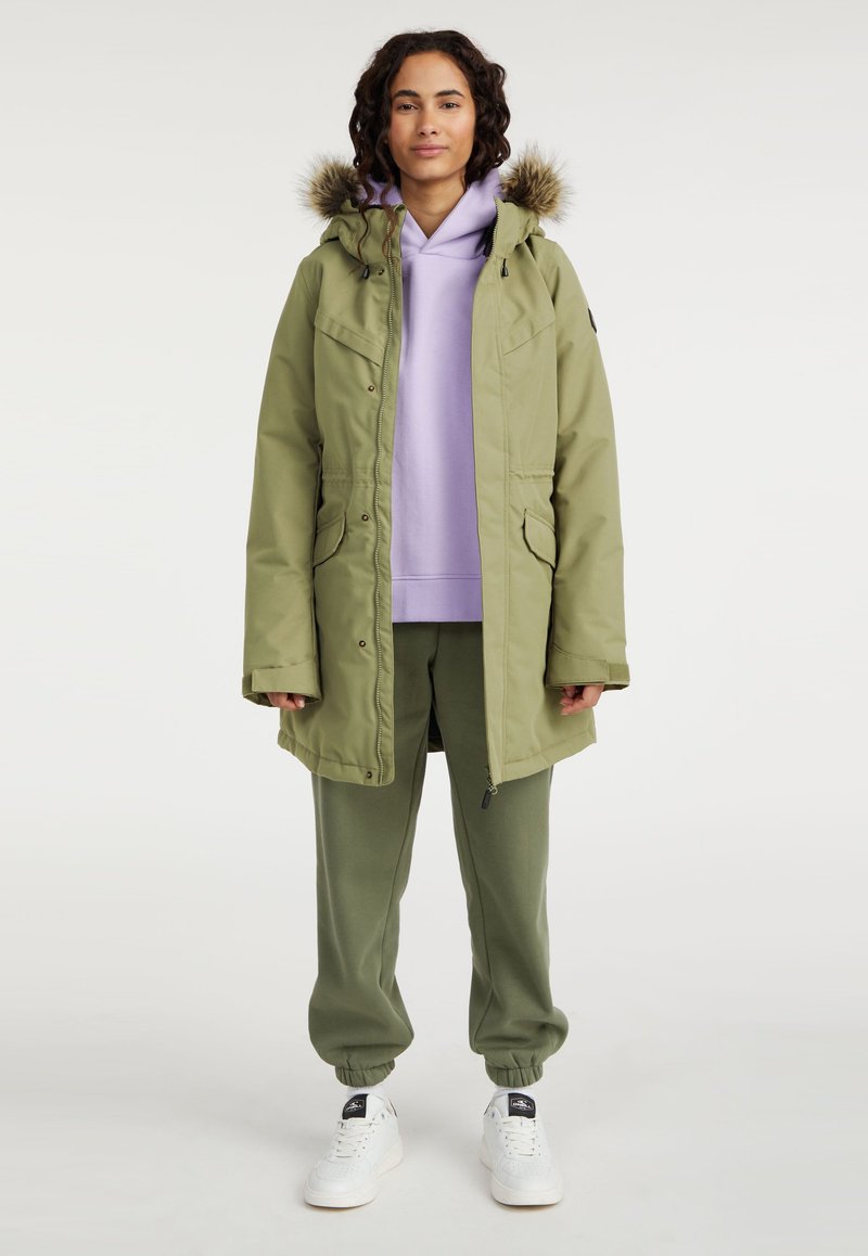 Olijfgroene parka met een gevoerde capuchon van bont, voorzien van drukknoopsluitingen en twee voorzakken, gedragen over een lichtpaarse hoodie en bijpassende groene joggingbroek.