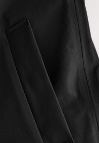 Gros plan d'un vêtement en tissu noir montrant des coutures détaillées et un rabat ou une poche rectangulaire.