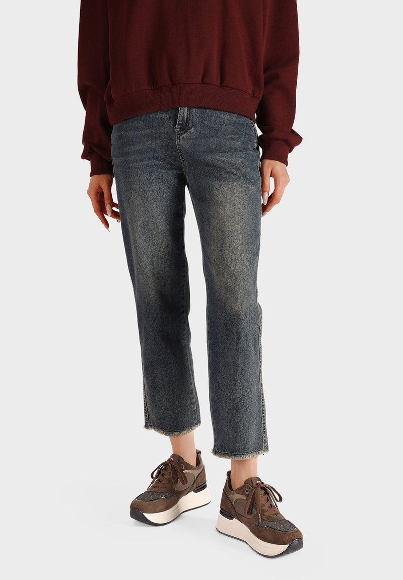 Jeans in denim blu scuro con orlo cropped e sfrangiato. Abbinati a sneakers marroni con dettagli testurizzati e suole color beige.