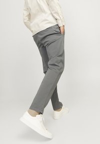 Personne portant un pantalon gris, des chaussettes beiges, des baskets blanches et une chemise à manches longues beige à rayures, les mains dans les poches, marchant avec une jambe levée.