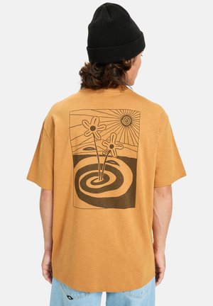 Person iført en sennepsgul t-shirt med en sort beanie, prydet med et sort abstrakt blomst- og soldesign på ryggen.