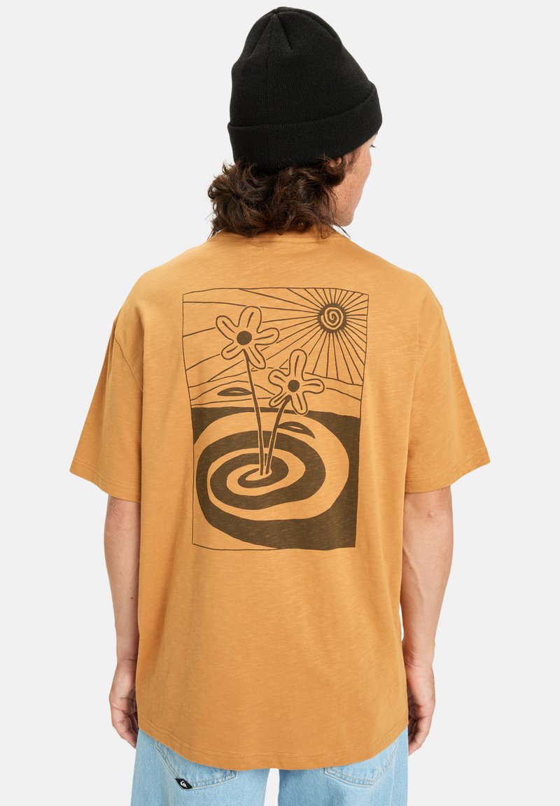 Person iført en sennepsgul t-shirt med en sort beanie, prydet med et sort abstrakt blomst- og soldesign på ryggen.