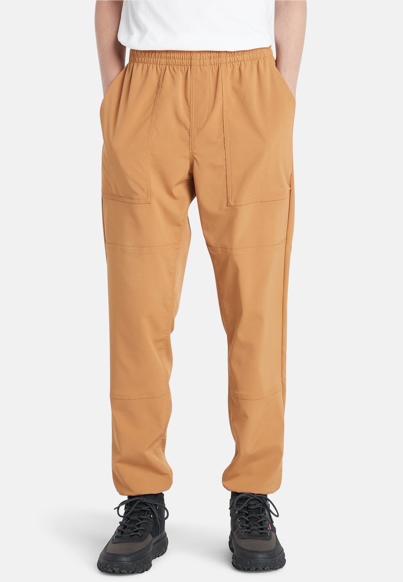 Tan cargobroeken met een elastische tailleband, voorzien van twee voorzakken en een losse pasvorm, gecombineerd met zwarte wandelschoenen.