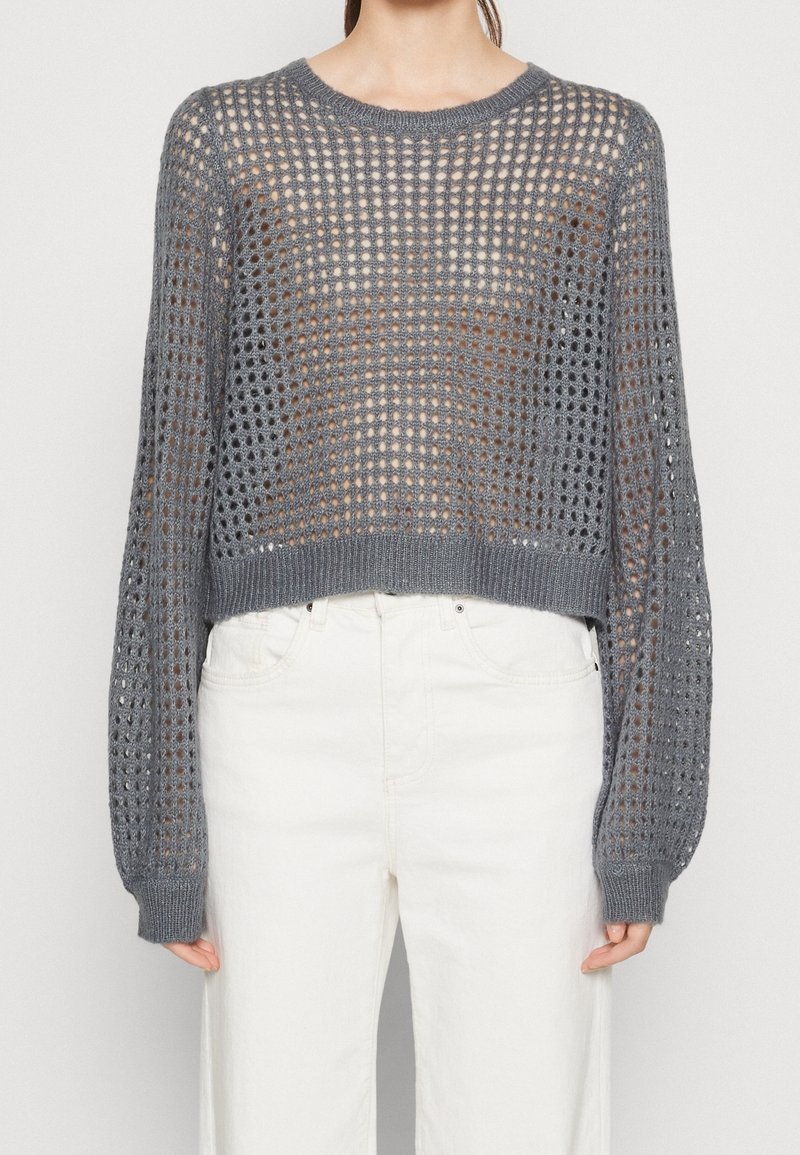 Femme portant un pull ample gris à maille ajourée sur un haut coloris peau, associé à un pantalon taille haute blanc, sur un fond uni.