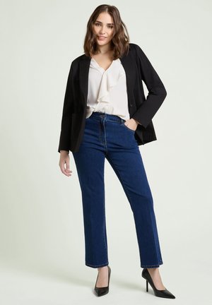 Ung kvinna i svart kavaj, vit blus, blå jeans och svarta högklackade skor står med ena handen i fickan och ler mot kameran.