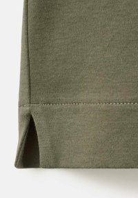 Marginea unui tricou verde măsliniu cu o despicătură laterală, realizată dintr-un material neted, texturat, cu cusături uniforme, care creează un finisaj curat.