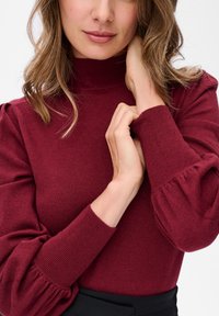 Kobieta o falujących, jasnobrązowych włosach, ubrana w burgundowy sweter z prążkowanej dzianiny z bufiastymi rękawami, delikatnie dotyka szyi i nadgarstka.