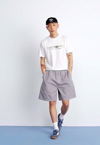 White cotton t-shirt with green print, gray shorts, black cap, and blue athletic shoes. Model stands against a plain background.