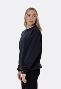 Felpa nera in tessuto morbido con vestibilità rilassata, spalle scese e polsini a coste. Design minimalista senza motivi o grafiche visibili.