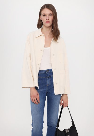 Lys beige cropped jakke i et mykt stoff, med skjortekrage, frontknapper og to sidelommer, kombinert med blå jeans.