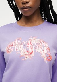 Jeune personne avec des tresses foncées portant un sweat lavande avec un motif floral rose et le texte "Versace Jeans Couture" sur la poitrine.