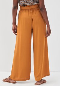 Personne portant un pantalon large taille haute de couleur orange brûlé avec un haut imprimé rentré, debout pieds nus sur un sol blanc contre un fond uni.