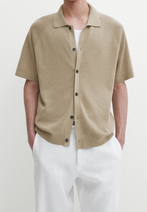 Homme portant une chemise tricotée beige à manches courtes et boutonnée par-dessus un sous-vêtement blanc et un pantalon blanc, les mains dans les poches.