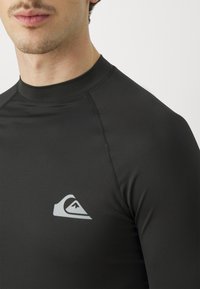 Czarny rash guard z długimi rękawami. Gładka tekstura, wysoki kołnierz oraz szwy płaskie. Wyróżnia się szarym logo na lewej piersi.