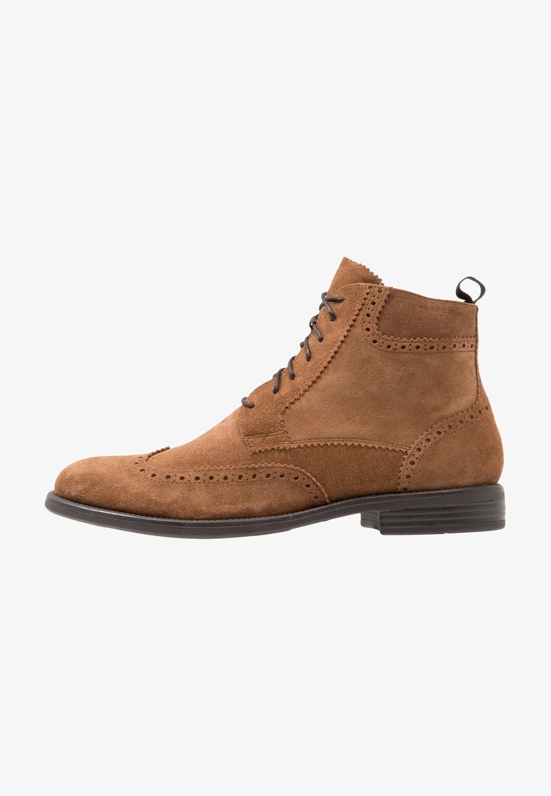 Bottine brogue en cuir suédé marron avec détails wingtip, lacets foncés, tirette et petit talon empilé sur fond blanc.