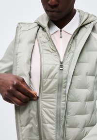 Personne portant une veste matelassée gris clair par-dessus un haut blanc zippé, tirant la fermeture éclair de la couche intérieure de la veste.