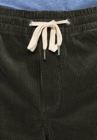 Gros plan sur un pantalon en velours côtelé vert foncé avec un cordon beige noué en nœud à la taille et une ceinture en tissu bleu visible au-dessus.