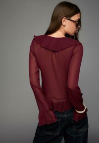 Woman wearing sheer burgundy blouse with ruffled collar and cuffs, paired with dark jeans, posing sideways against gray background.