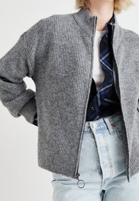 Szary dzianinowy kardigan o prążkowanej fakturze, z wysokim kołnierzem i zamkiem z przodu. Nałożony na niebieską koszulę w kratę, zestawiony z spodniami z wysokim stanem.