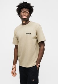 Χακί βαμβακερό T-shirt με μαύρο λογότυπο "Dickies" στο στήθος. Η μπλούζα έχει άνετη εφαρμογή και κοντά μανίκια. Φορεμένο με μαύρα cargo σορτς.