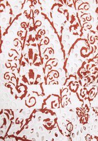 Tissu blanc avec broderie florale et volutes complexes en relief couleur rouille rouge et petits œillets partout.