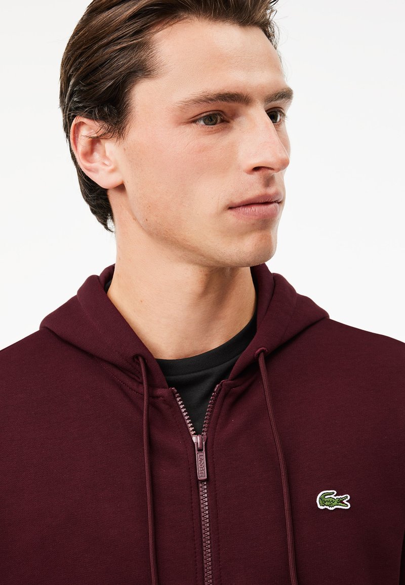 Un sweat à capuche zippé bordeaux en coton doux, avec un logo de crocodile vert sur la poitrine gauche et des cordons de serrage sur la capuche.