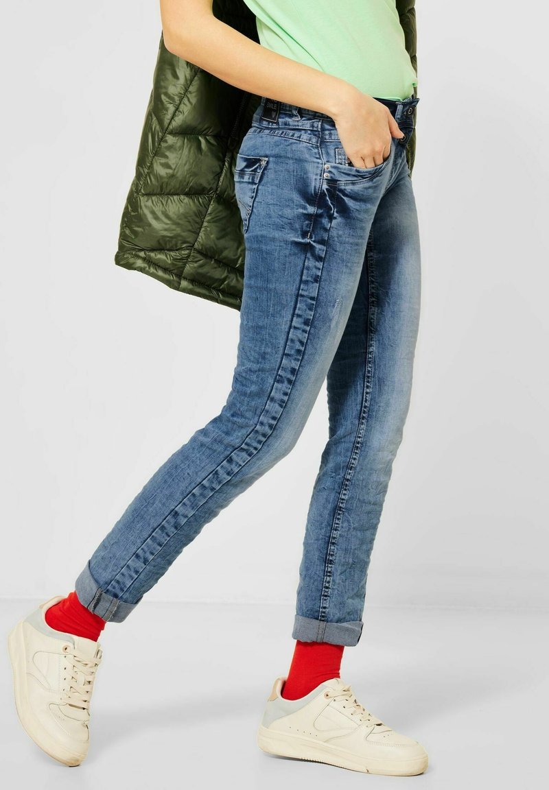 Personne portant un jean bleu retroussé, des chaussettes rouges, des baskets blanches, un haut vert clair et tenant une doudoune vert foncé sur l'épaule.