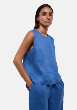 Femme aux longs cheveux foncés portant un haut sans manches en lin bleu et un pantalon en lin bleu assorti, les mains dans les poches, debout devant un fond blanc.