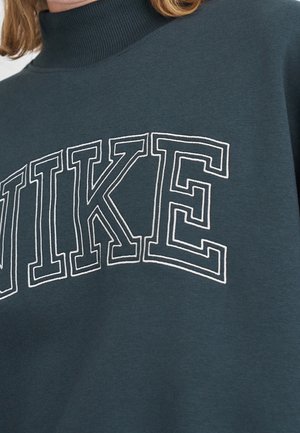 Nærbillede af mørkegrøn sweatshirt med hvide markerede bogstaver, der staver "NIKE" henover brystet og ribbet høj krave.
