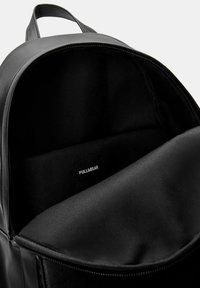 Zaino in pelle nera aperto per mostrare l'interno in tessuto liscio con una piccola etichetta del marchio Pull&Bear all'interno del compartimento principale.
